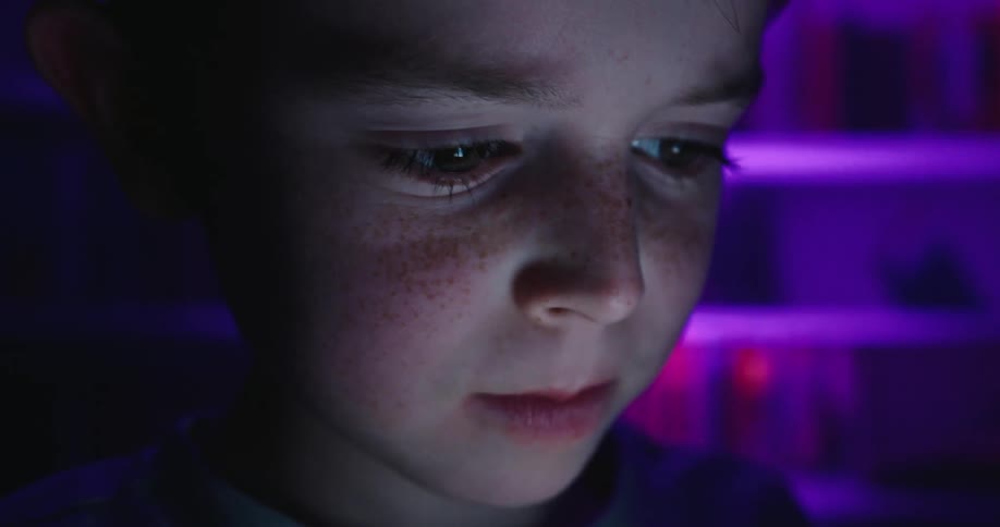 A young boy illuminated by purple light smiles while looking at a smartphone in a dark room
