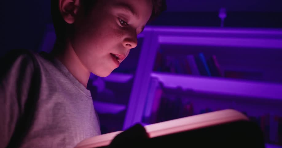 A young boys face is illuminated with wonder as he reads from a magical book that glows warmly in his hands while purple light fills the room behind him