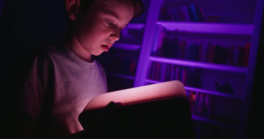 A child indoors holds a book that glows as he read from it while it glow softly illuminating his face as he gazes at it with curiosity