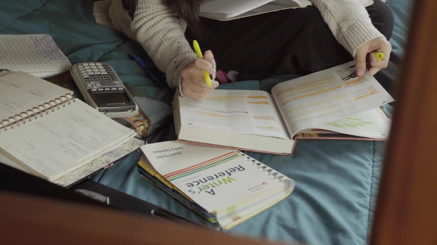A woman sits on a bed studying with books and a calculator