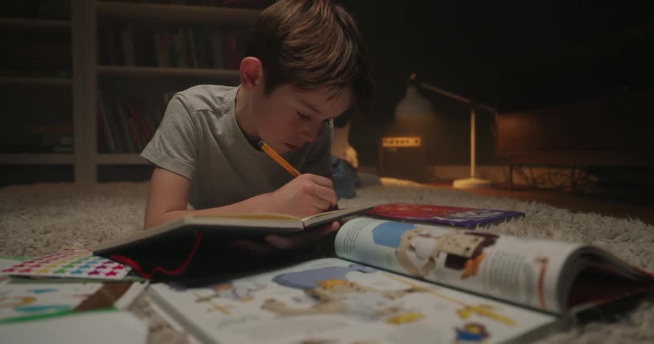 Boy lying on the floor at night with books writes in a journal