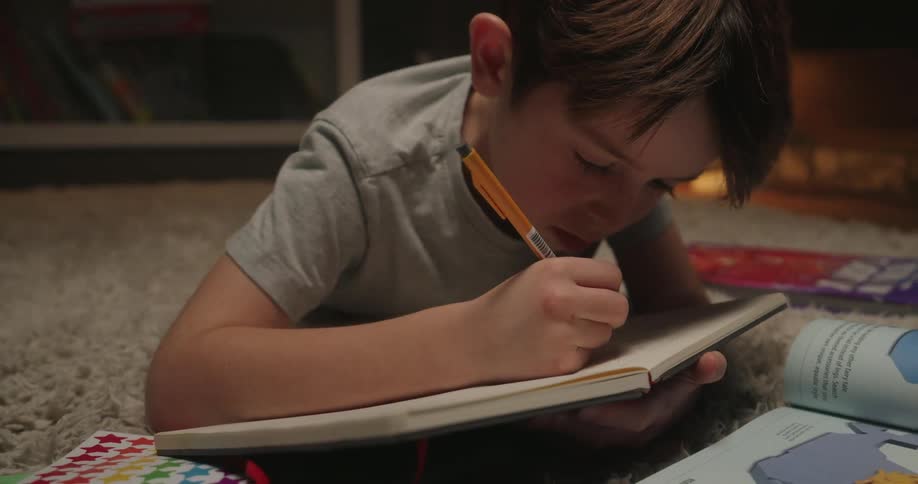 A young boy lays on the floor while writing on a notebook page