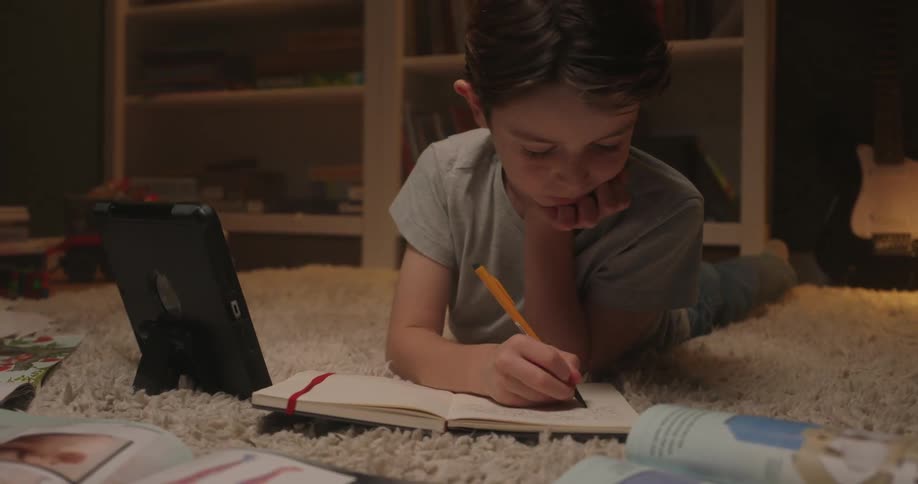 A young boy is laying on a rug in a dimly lit room writing in a notebook while looking at a standing tablet