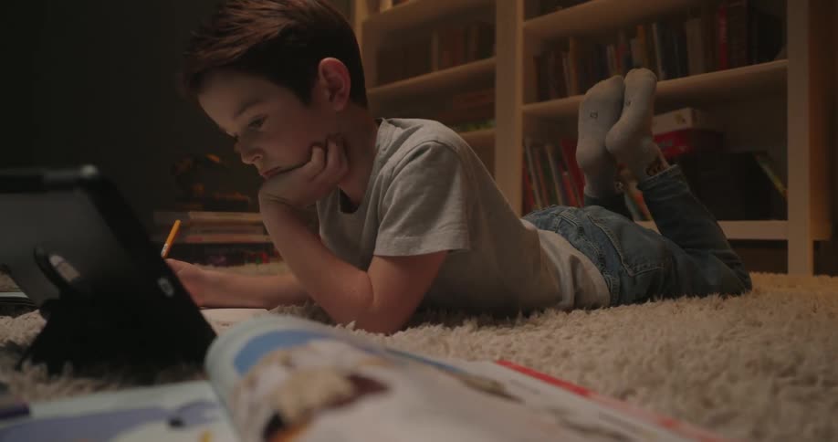 A young boy lies on a carpet using a tablet and writing in a notebook near a bookshelf