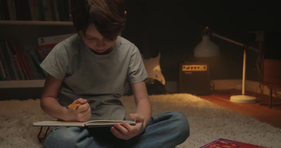 A boy is sitting in a dimly lit room writing in a notebook on a large rum