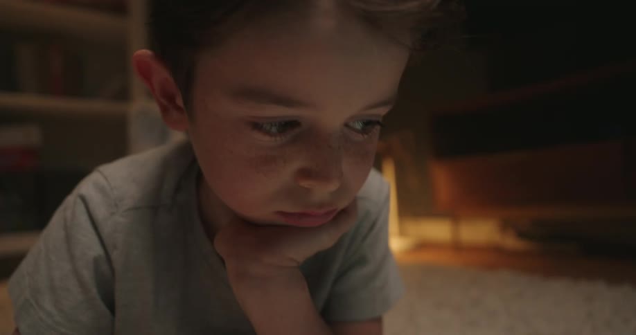 A close view of a thoughtful young boy lying on a carpeted floor writing in a notebook with a yellow pencil next to a tablet while resting his chin on his hand