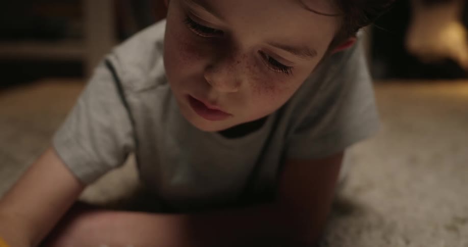 A close view of a young boy with freckles lying on a shaggy rug concentrating while writing in a notebook with a yellow pencil in a dimly lit room