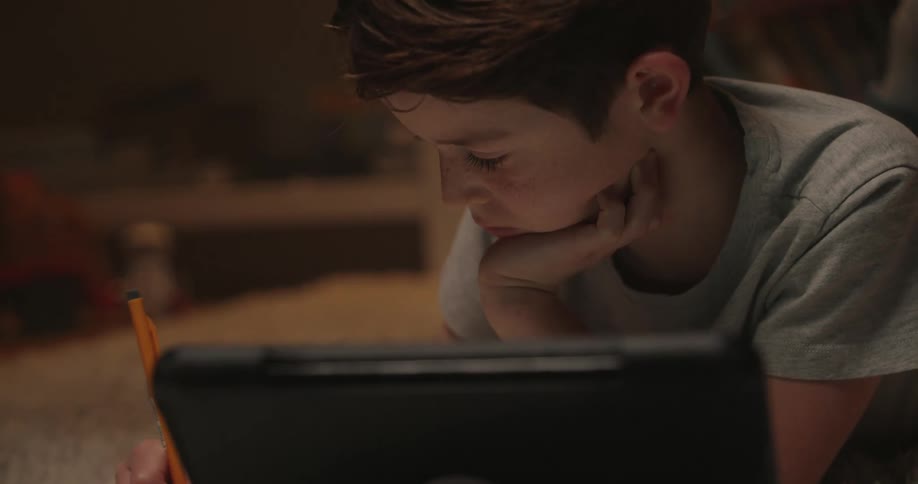A focused male child with freckles is concentrating on a digital device in a dimly lit room