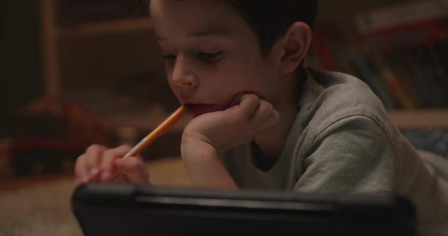 This video shows a closeup of a young boy with dark hair and freckles lying down in a dimly lit room likely at nighthe is focused on a tablet in front of him resting his head on his hand and briefly chewing on the end of an orange pencil before retu