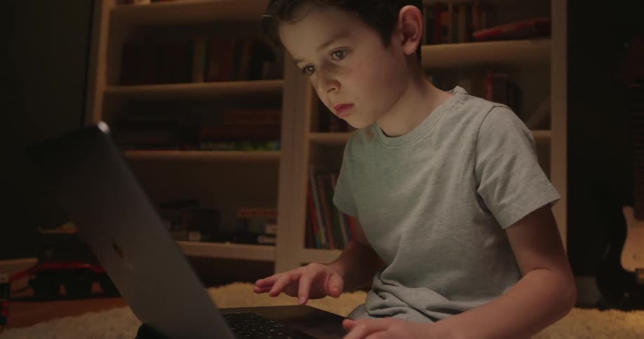 A young boy wearing a gray shirt sits on a rug in a dimly lit room with bookshelves and a guitar intensely using a laptop computer