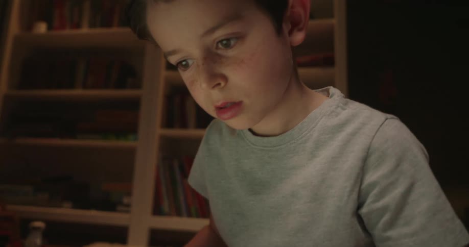 A young boy wearing a gray shirt sits on the floor in a dim room with bookshelves and a guitar intensely focusing on the laptop screen in front of him