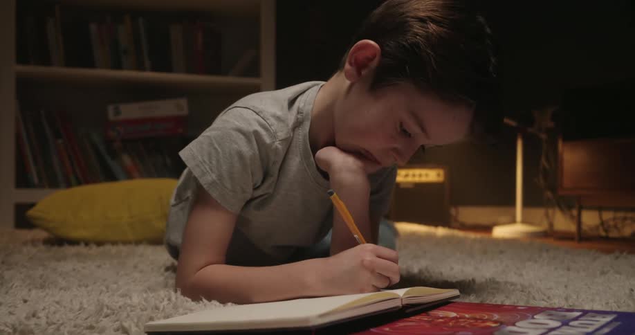 Child draws and writes in a notebook while papers and books are spread across the rug