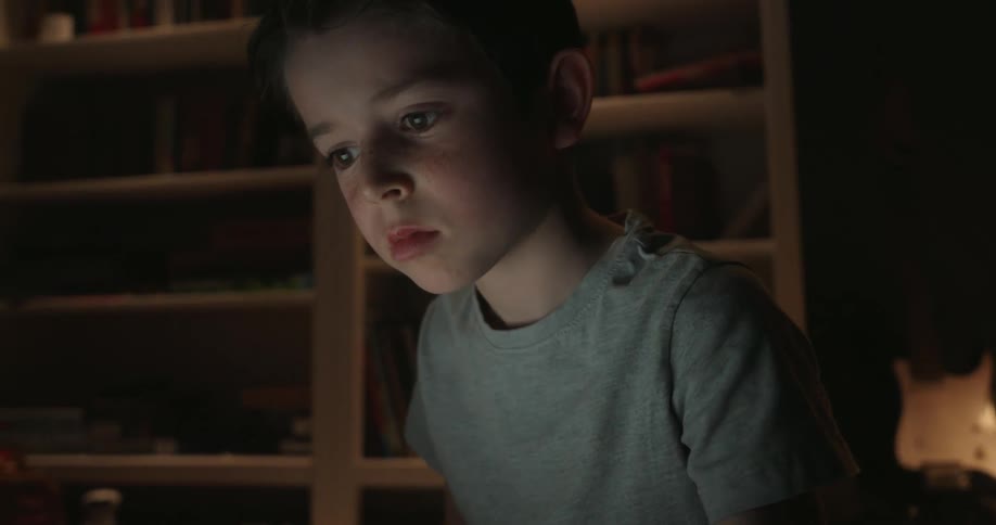 A close view of a young boy in a gray shirt sitting in a dark room with bookshelves focused on typing on a laptop whose light illuminates his face