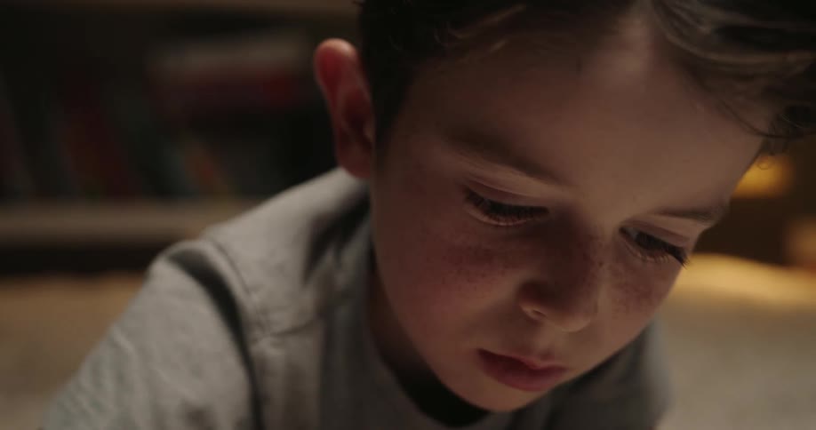 A young boy lies on a plush rug writing in a notebook near a softly lit bookshelf indoors