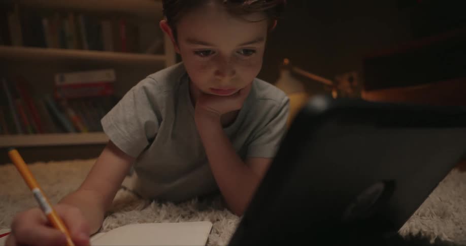 A young boy with freckles is seen lying on a rug in a dimly lit room diligently writing in a notebook with an orange pencil he frequently looks up to check information on a tablet propped up in front of him suggesting he is working on a homework assign