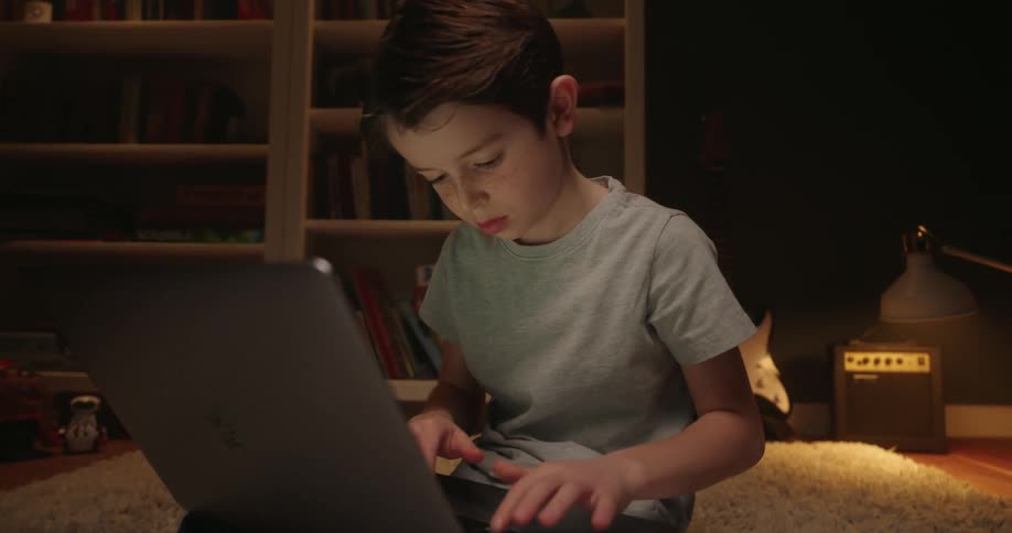 A young boy in a gray tshirt sits on a rug using a laptop in a dimly lit room with bookshelves a guitar and an amplifier visible in the background