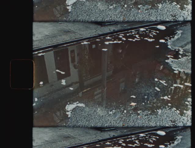 The video is a closeup topdown shot of a large puddle on an asphalt surface possibly near a curbthe water reflects the muted image of nearby buildings and the surrounding concreteasphalt and the clip is presented with a grainy vintage film effe