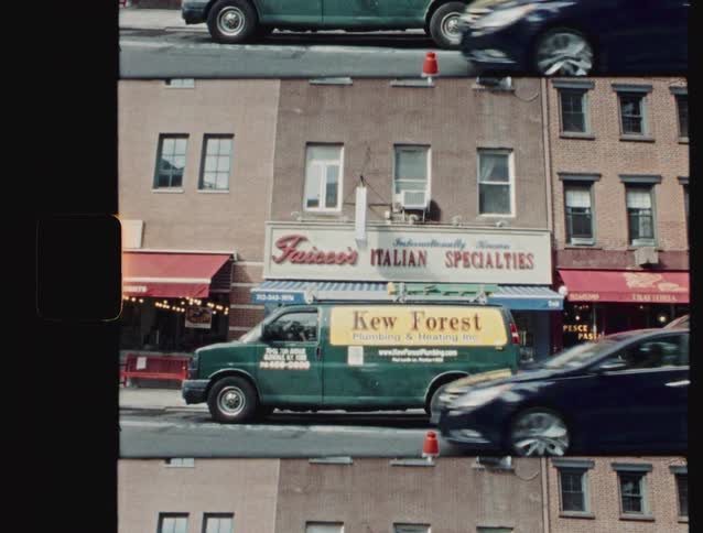 Captured on vintage film with visible sprocket holes a green plumbing van sits parked in front of an italian specialty shop while traffic blurs past on the busy city street