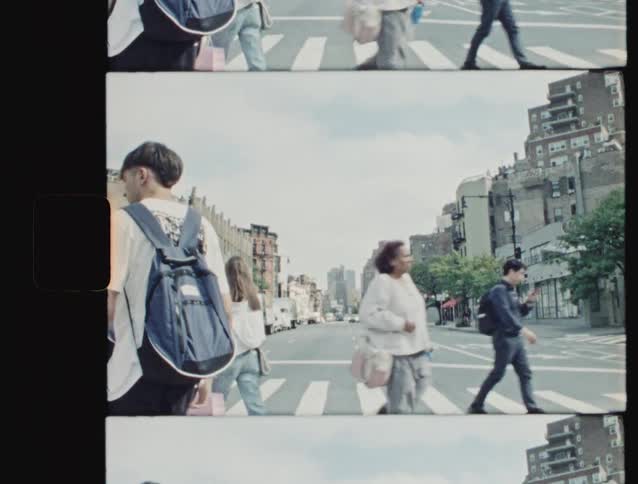 Pedestrians some with pets or a stroller cross at a crosswalk a distance from a skyline on a cloudy day