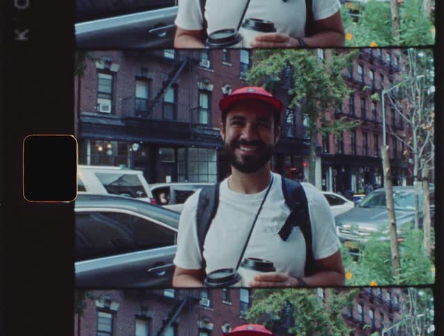 The video shows a smiling man standing on a city street possibly in new york holding two disposable coffee cupshe is wearing a white tshirt a red baseball cap and a black backpack with what looks like a camera strap around his neck and the vide