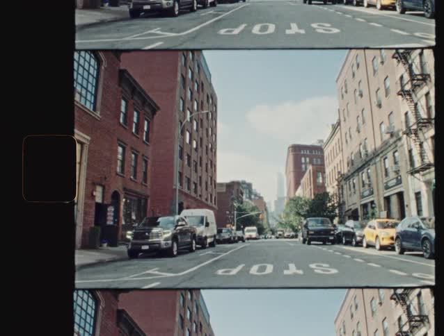 A series of cars are parked along the side of the street while tall apartment buildings line the road the midday sun casts clear light across the scene highlighting the urban setting