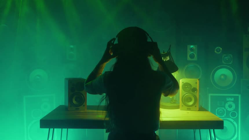 Person standing in front of a table in a room with audio speakers puts on headphones as colored lights flash