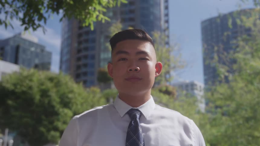Man in a dress shirt and tie stands outdoors with buildings in the background