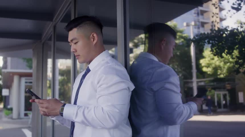 Man standing outside next to a building texting on his phone with his reflection on the window