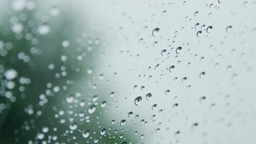 Closeup of a window covered in distinct water droplets with the texture of the wet glass being the focus while a blurred outoffocus background of green foliage and a light sky remains visible beyond