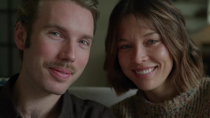 Young adult man and young adult woman sitting down next to each other on a couch and smiling indoors during day time