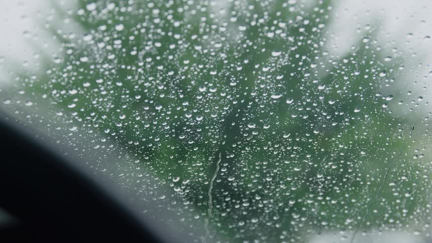 A closeup shot focuses on numerous small rain droplets covering a window or windshield with a single stream of water slowly trailing down the center and a blurred green background visible