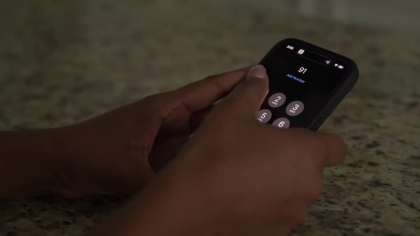 A closeup shot shows a persons hands holding a black smartphone over a granite countertop quickly dialing the digits 9 and 1 twice on the illuminated keypad interface