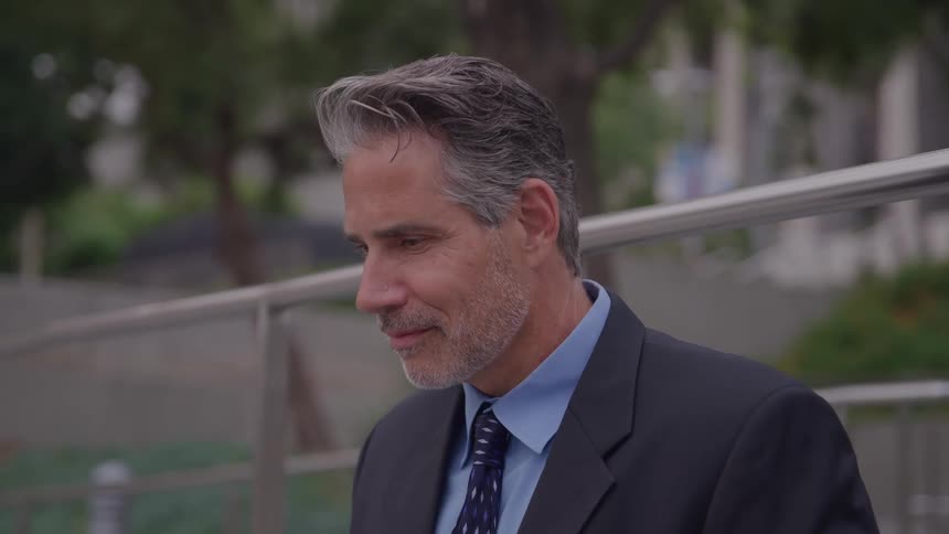 The video is a medium closeup shot of a mature man in professional attire looking down and smiling slightlythe man has saltandpepper hair is wearing a dark suit jacket over a light blue collared shirt and a patterned dark tiehe is outdoors in