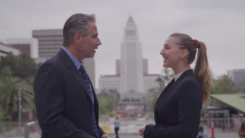 The video shows a man and a woman in business attire having an animated conversation outdoors in a city plazathe man with gray hair listens intently to the woman who has her hair in a ponytail and is actively gesturing including pointing up in the