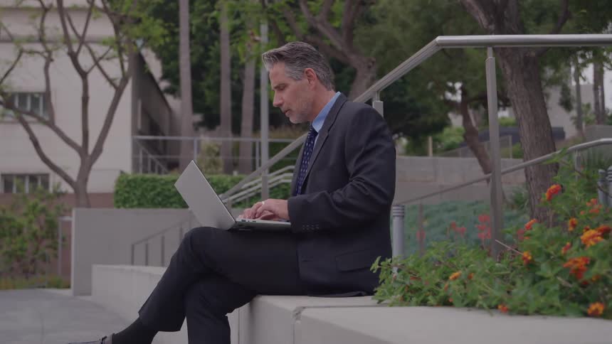 A middleaged man in a dark suit sits on a concrete ledge in an urban outdoor setting focused on typing on a silver laptop resting on his knees with a bush of orange and red flowers in the foreground