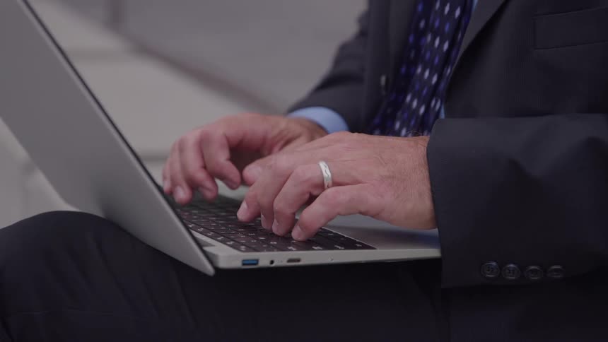 Man in a suit types on a laptop resting on his lap