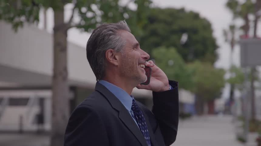 A sideprofile shot captures a middleaged man in a dark business suit standing outside holding a cell phone to his ear and smiling broadly while engaged in a conversation