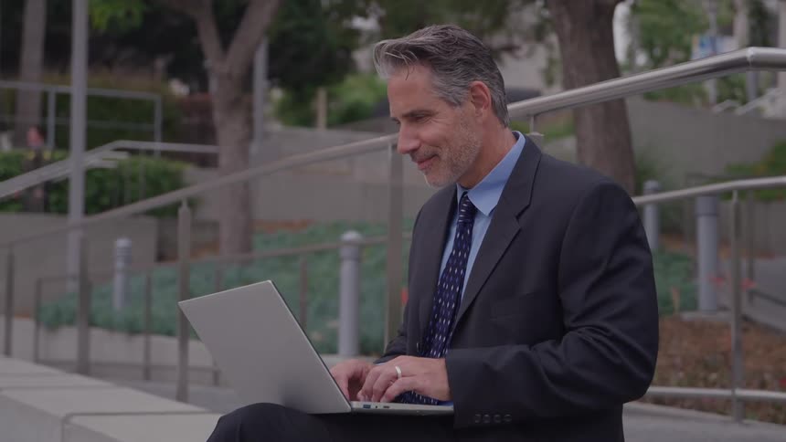 Man in a suit types on a laptop while sitting in an outdoor urban area