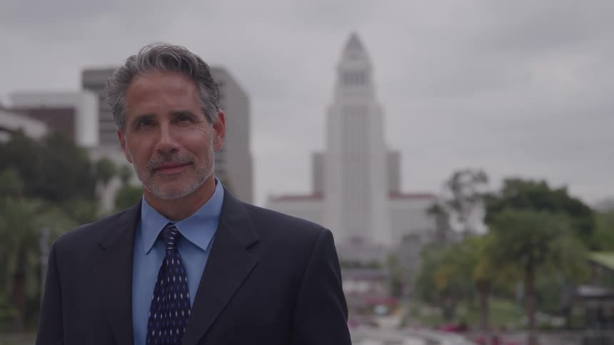 A confident middleaged businessman with graying hair wearing a dark suit smiles directly at the camera while standing outdoors with a tall city hall tower and surrounding urban buildings blurred in the background