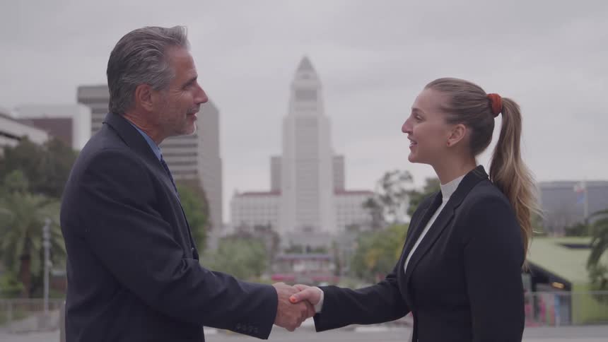 A businessman and businesswoman shake hands and smile in front of los angeles city hall