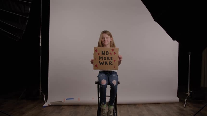 A young girl sits on a stool in a studio holding a cardboard sign that reads no more war