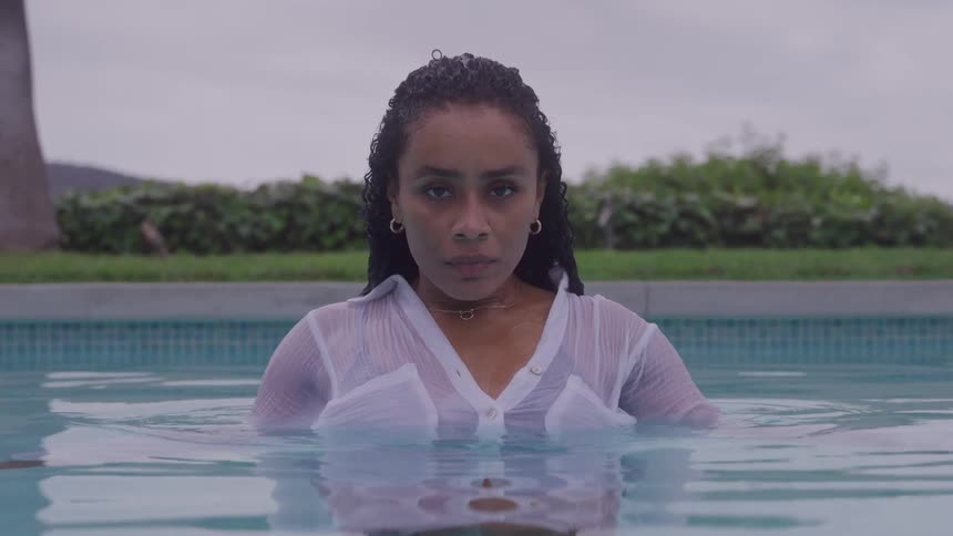 A woman with wet braided hair slowly emerges from the water in a swimming pool wearing a soaked white shirt and maintaining intense eye contact with the viewer