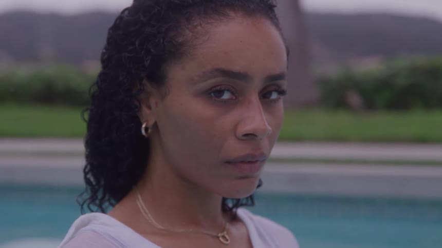 Closeup side profile of a woman with curly hair looking away then turning her gaze to meet the camera with a serious expression positioned outdoors next to a bright blue swimming pool