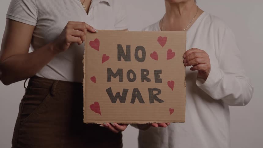 Two women hold a handmade cardboard sign decorated with red hearts and the message no more war written in black marker against a plain background