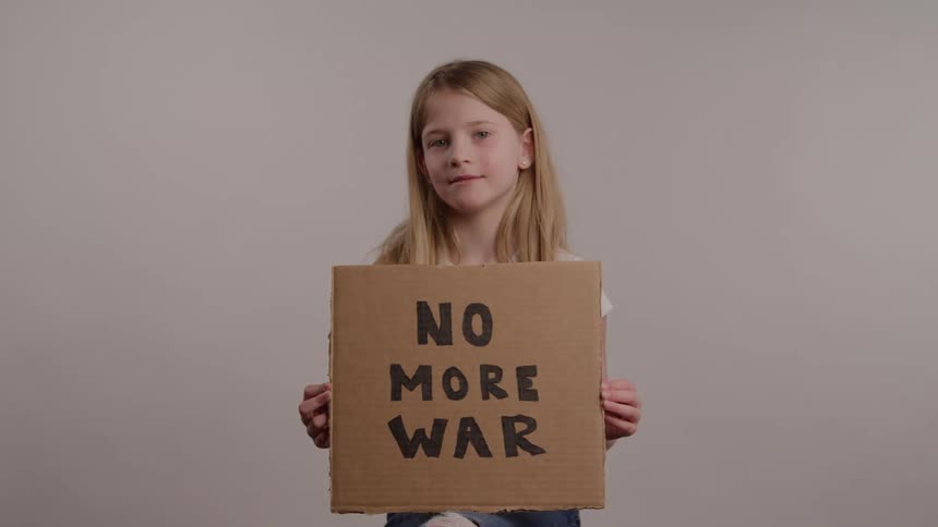 A young girl with long blonde hair stands in front of a neutral background holding a square piece of cardboard sign that boldly reads no more war in black lettering