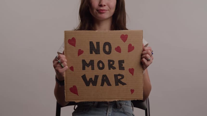 Seated woman holds a cardboard sign with the phrase no more war decorated with red hearts