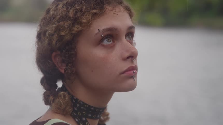 A close view of a young woman with curly brown hair and facial piercings looking upwards against a soft gray background of water and sky