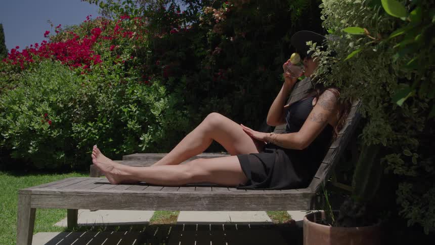 Woman lying on a lounge chair drinks a beverage outdoors surrounded by plants