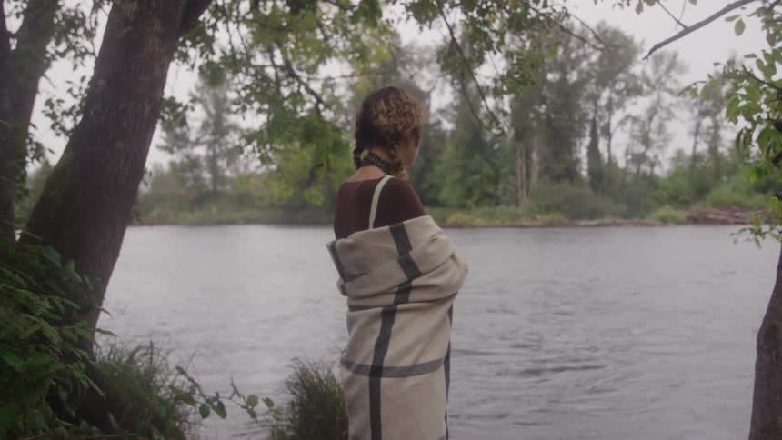 A medium shot from behind shows a woman wrapped tightly in a thick patterned blanket standing by the edge of a wide river and gazing out at the lush forested banks on the opposite side