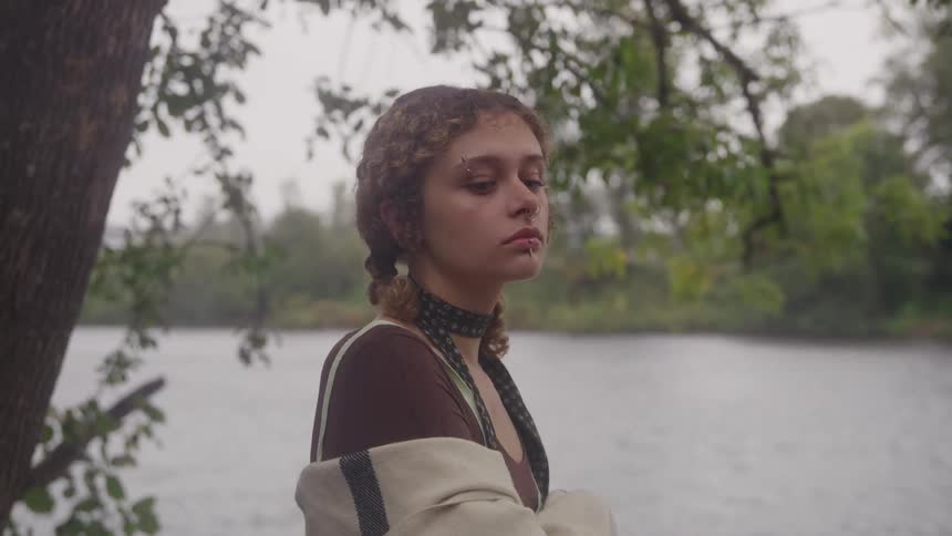 A pensive young woman wrapped in a shawl stands by a river on an overcast day